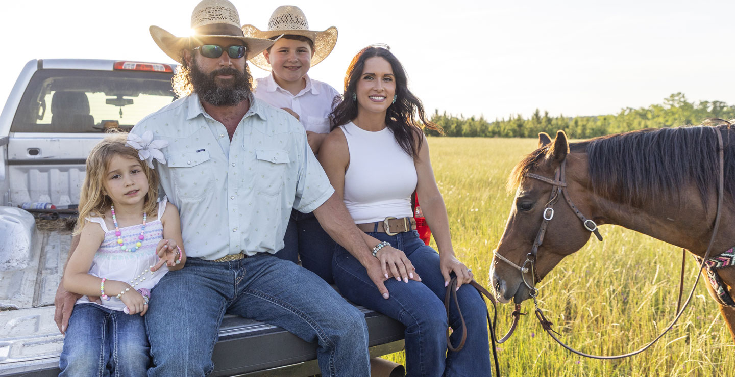 The Smith family sitting in the back of a truck, smiling, while Kip Smith holds the lead rope to a brown horse standing beside the truck. 